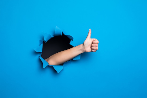 Close up of woman hand with thumbs up gesture through blue hole in paper wall. Free Photo