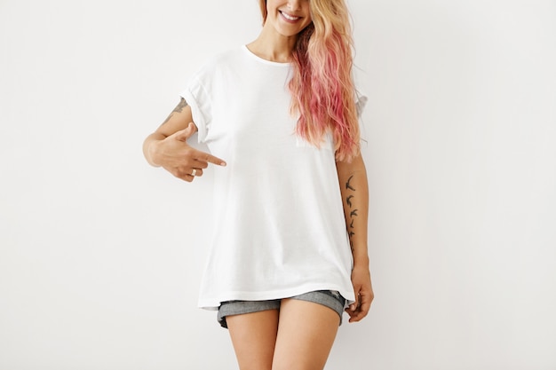 服 デザイン スタイル ファッション 広告 ピンクの髪のハイライトと入れ墨の腕の笑顔 空白のtシャツで指を指している認識できないスリムな流行に敏感な 女性の肖像画 無料の写真
