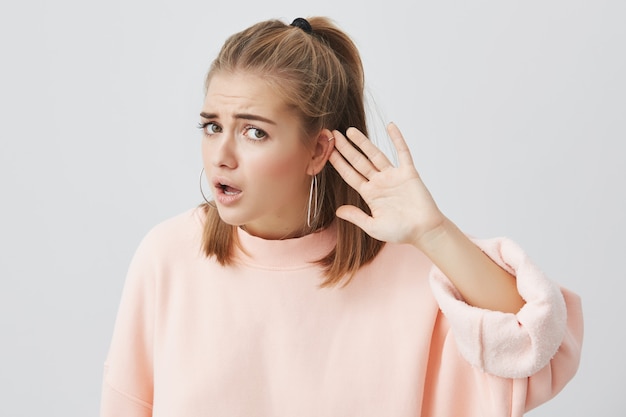 Clueless caucasian woman in pink sweater looking  with confused and puzzled expression, frowning, holding hand behind ear like she doesn`t hear what she is told to do, ferusing to do that Free Photo