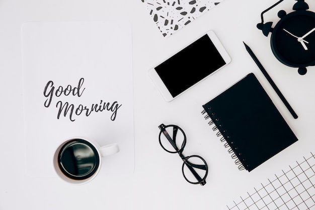 Coffee cup over paper with good morning text; cellphone; alarm clock and stationeries on white desk Free Photo