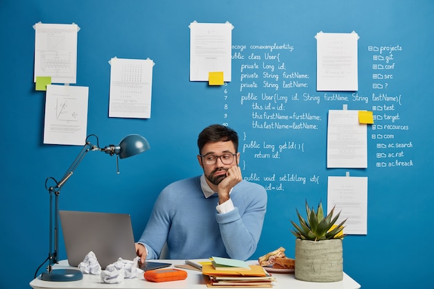 Concentrated bearded web developer improves new website version, sits at white table, loaded with notepads, snack, cup of tea and potted plant, looks sadly at problem of project, leans at hand Free Photo