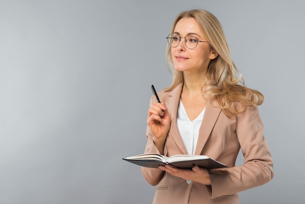 Confident smiling blonde young woman holding pen and diary in hand against gray background Free Photo