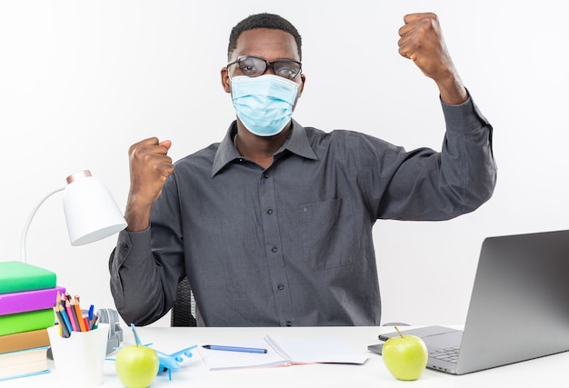 Confident young afro-american student in optical glasses wearing medical mask sitting at desk with school tools raising fists up Free Photo