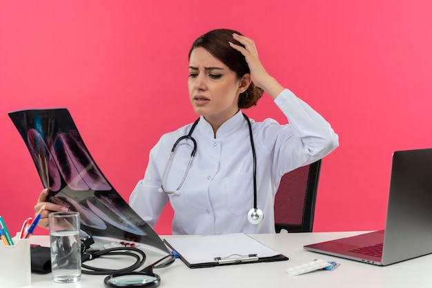 コンピューターを保持しているとコピースペースとピンクの壁に頭に手を置くx線を見て机の仕事に聴診器で医療ローブを着て混乱している若い女性医師 無料の写真