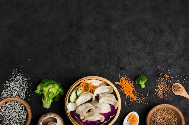 Cooked dumplings inside the bamboo steamer with eggs; broccoli; sesame and coriander seeds on black texture background Free Photo
