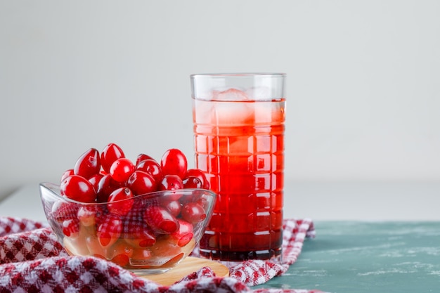 Cornel berries with drink, cutting board, picnic cloth in a bowl on plaster and white, side view. Free Photo