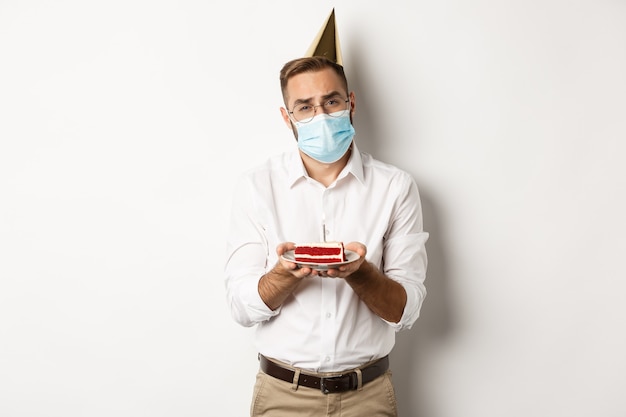 コロナウイルス 検疫および休日 悲しい男はバースデーケーキからろうそくを吹き消すことができない フェイスマスクを着用して不平を言って立っている 無料の写真