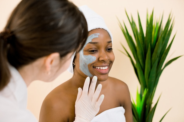 Premium Photo Cosmetologist applying mask onto face of african Premium Photo Cosmetologist applying mask onto face of african