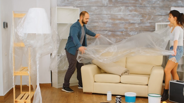 Couple wrapping sofa in plastic foil for protection while they are renovate living room. home renovation, construction, painting work. Free Photo