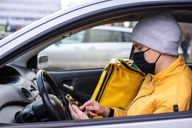 黒の医療用マスクを付けた車の宅配便は彼の電話にあり 配達用バックパックは座席にあります フードデリバリーサービス 無料の写真