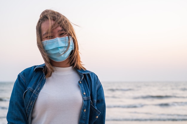 Covid-19 concept, asian young woman wearing a hygiene protective mask over her face outdoor with sunset at the beach Premium Photo