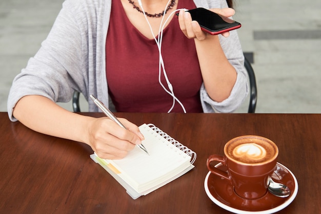 同僚へのボイスメッセージを録音し カフェのテーブルに座っているときにプランナーでメモを取る女性起業家のトリミングされた画像 プレミアム写真