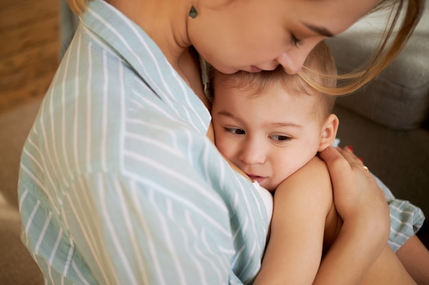 優しい思いやりのある若い母親が眠そうな赤ちゃんを腕に抱きしめ 彼に歌を歌い 額にキスをしている様子のトリミングされた画像 1歳の小さな子供を抱き締め 腹部のけいれんを和らげようとしているお母さん 無料の写真