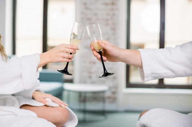 シャンパンのグラスを押しながら乾杯する白いバスローブを着た2人の手の写真をトリミングしました 完全なシャンパングラスを持つ男と女の手 プレミアム写真