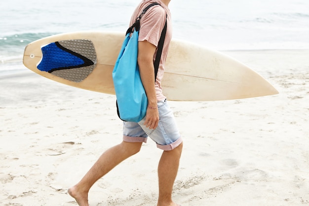 Cropped shot of barefooted man with blue bag carrying white surfboard ...