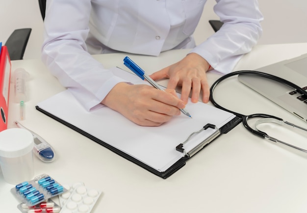 Cropped shot of female doctor hands working at desk with medical tools and laptop writing prescription on clipboard Free Photo