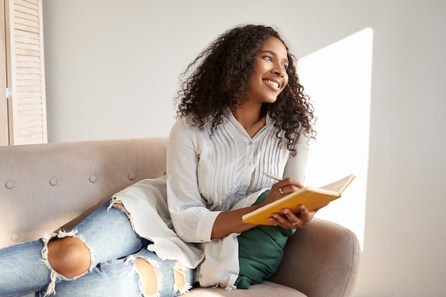 Cute adorable afro american student girl with black voluminous hair enjoying leisure time after college, lying on couch in stylish ripped jeans and blouse, sharing thoughts and ideas in her diary Free Photo
