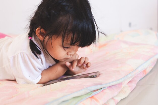 彼女の部屋で彼女のベッドに横になっているスマートフォンを弾くかわいいアジアの赤ちゃん女の子 プレミアム写真
