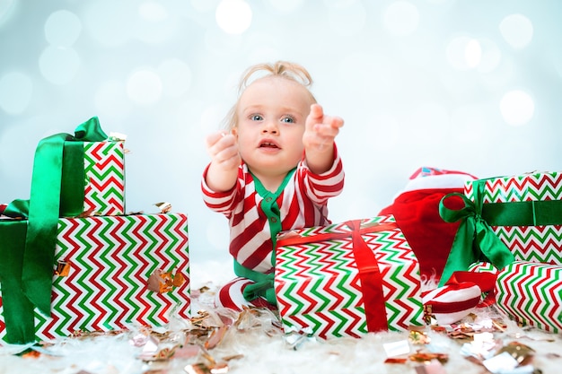 クリスマスの背景にポーズをとってサンタ帽子に近い1歳のかわいい赤ちゃん女の子 クリスマスボールが付いている床に座っています 連休シーズン 無料の写真