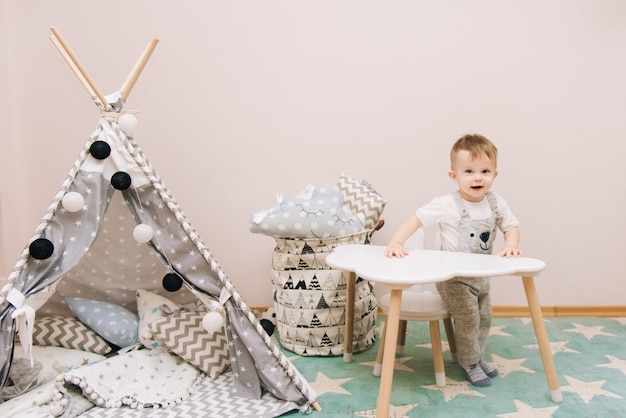 白 グレー ブルーの色調で子供部屋のテーブルに座っているかわいい赤ちゃん ティーピーとおもちゃの袋の近く プレミアム写真