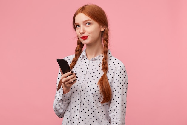 手に携帯電話を持っているかわいい美しい赤毛の女の子は 右上隅にコケティッシュに不思議に見えます ピンクで隔離されたメッセージで彼女のボーイフレンドに何を書くべきかを考えています 無料の写真