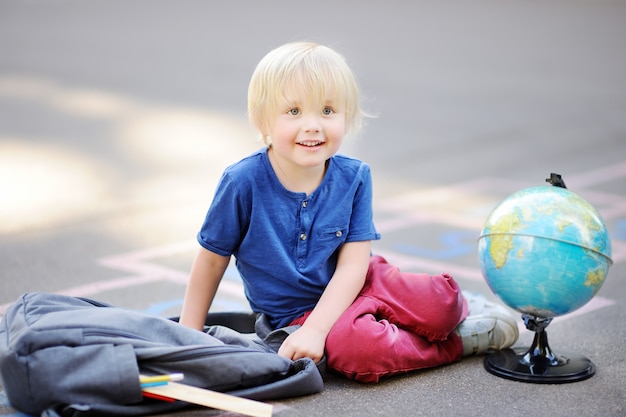 Cute blond boy doing homework sitting on school yard after school with bags laying near. Premium Photo