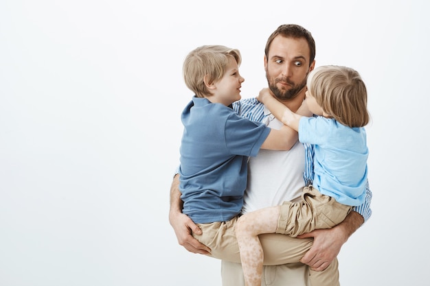 かわいい面白い白人の父親が2人の若い息子を腕の中で保持し 年下の少年を見つめて顔を作り 素晴らしい子供たちがいる間に前向きで幸せになる 無料の写真