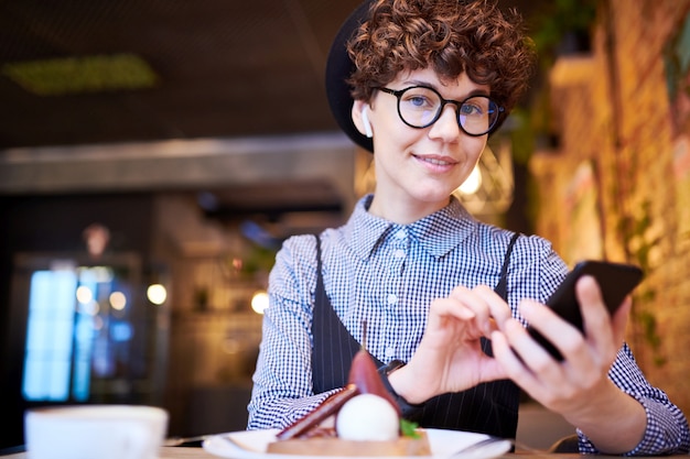 プレミアム写真 スマートカジュアル 帽子 Airpods 眼鏡でかわいい女の子がカフェに座っている間スマートフォンでプレイリストを見て