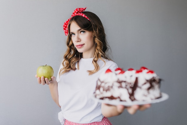 カロリーを考えてケーキを持っている暗いウェーブのかかった髪のかわいい女の子 リンゴとクリーミーなチョコレートパイを持つかっこいい若い女性の屋内写真 無料の写真