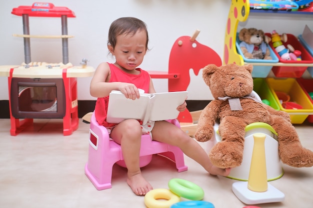 かわいい小さなアジアの18ヶ月の幼児の女の赤ちゃんがトイレに座って おもちゃとテディベアと一緒に自宅のプレイルームで本を読んでいます プレミアム写真