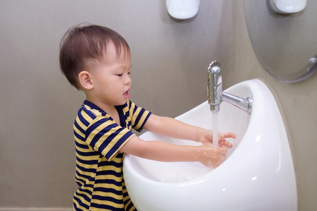 かわいい小さなアジアの2歳の幼児の赤ちゃん男の子子供一人で白いシンクと手を洗って公衆トイレの蛇口から水滴 子供用バスルーム 衛生 衛生概念 プレミアム写真