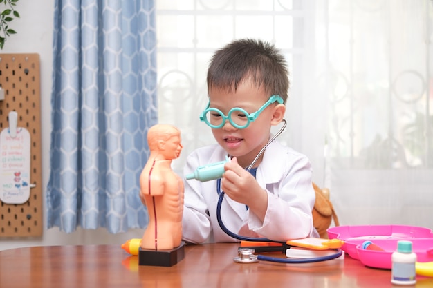 医者の制服を着たかわいいアジア4歳の学校の男の子が自宅で医者を再生 子供が聴診器を身に着けていると解剖学的身体器官モデルで遊ぶ プレミアム写真