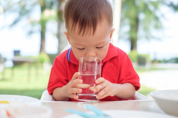 かわいいアジアの幼児男の子の子供がビーチリゾートのレストランで一人で水のガラスを保持 飲むガラスに座って プレミアム写真