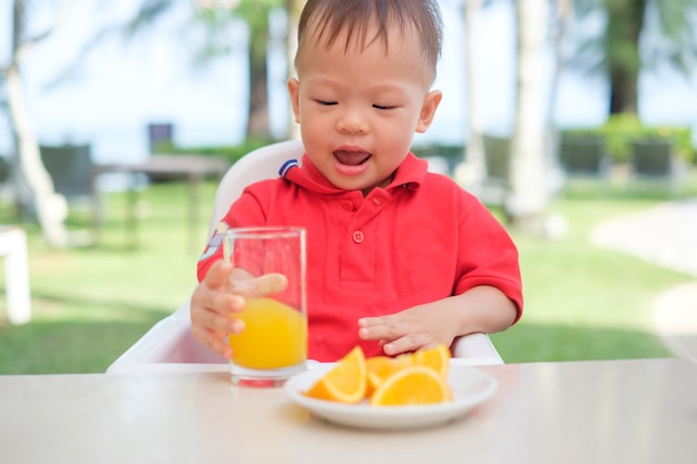朝食時にガラスでおいしいオレンジジュースの冷たい飲み物を保持 飲む高い椅子に座っているかわいい小さなアジア幼児男の子 プレミアム写真