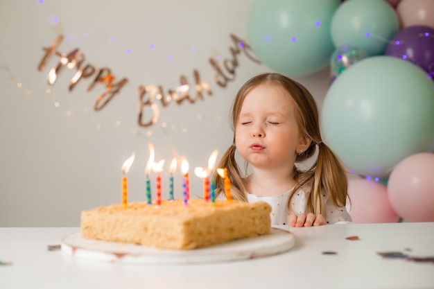 かわいい女の子は 風船を背景に自宅でバースデーケーキにろうそくを吹きます 子供の誕生日 プレミアム写真
