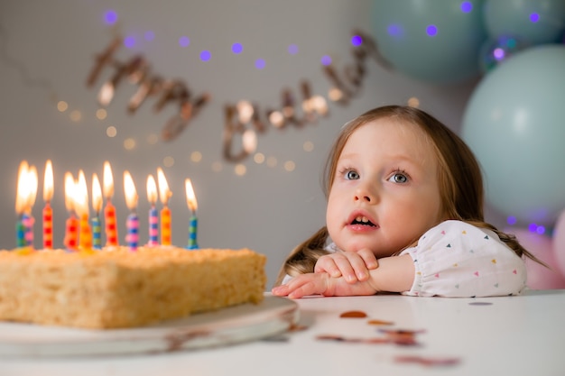 かわいい女の子は 風船を背景に自宅でバースデーケーキにろうそくを吹きます 子供の誕生日 プレミアム写真