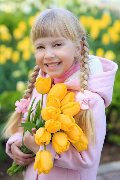 美しい花の背景に黄色いチューリップの花束を持っているかわいい女の子 ピンクのコートを着た女の子 プレミアム写真