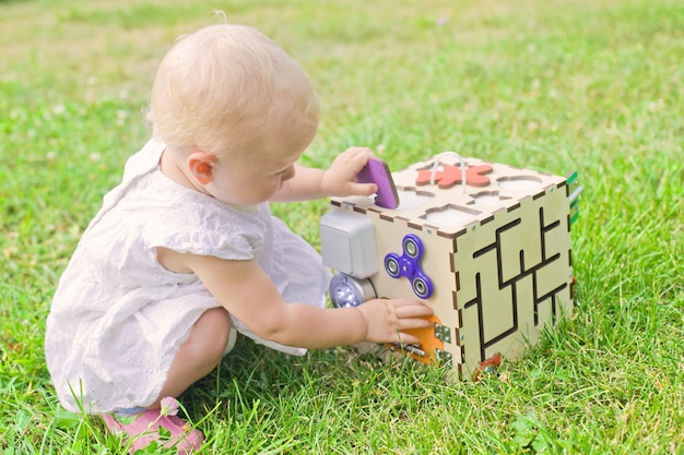 かわいい女の子は屋外の緑の芝生でビジボードで遊んでいます 幼児向けの教育玩具 女の子は ボードのキューブへの扉を開きました プレミアム写真
