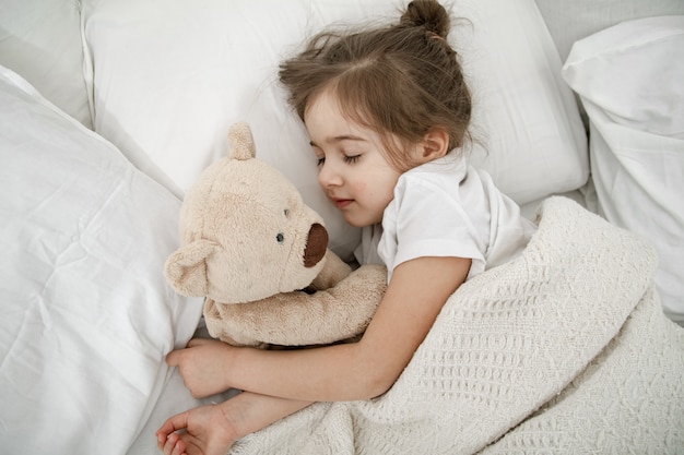 A cute little girl is sleeping in a bed with a teddy bear toy. Free Photo