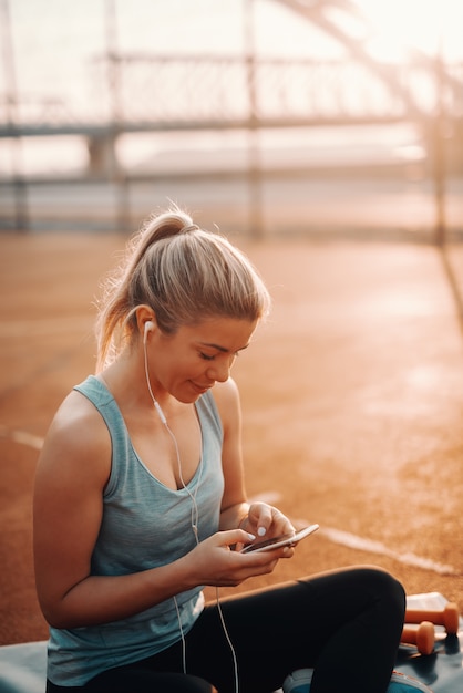 かわいい笑顔白人金髪スポーティな女性の耳にイヤホンでコートに座って スマートフォンを使用しています プレミアム写真