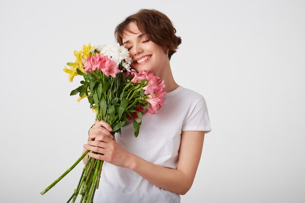 色とりどりの花の花束を持って 匂いを楽しんで 目を閉じて広く笑って 白い背景の上に立って 白い空白のtシャツを着たかわいい若い短い髪の少女 無料の写真