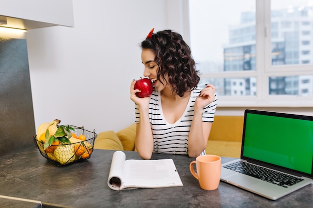モダンなアパートメントのテーブルに巻き毛を持つかわいい若い女性 赤いリンゴを食べて 目を閉じて笑みを浮かべて 自宅で職場 緑色の画面でノートパソコン 休憩 無料の写真