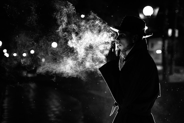 帽子をかぶった男の暗いシルエットノワールのスタイルで夜の街で雨の中でタバコを吸う プレミアム写真