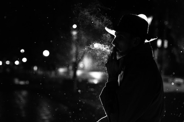 帽子をかぶった男の暗いシルエットノワールのスタイルで夜の街で雨の中でタバコを吸う プレミアム写真