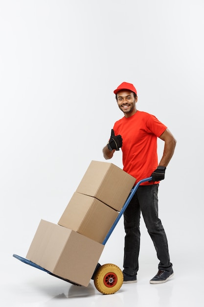 Delivery concept - portrait of handsome african american delivery man or courier pushing hand truck with stack of boxes. isolated on grey studio background. copy space. Free Photo