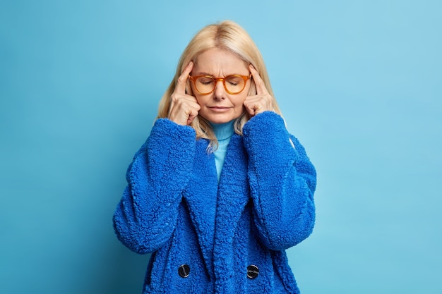Depressed blonde forty years old woman keeps fingers on temples thinks deeply or has headache closes eyes as tries to concentrate wears winter coat. Free Photo