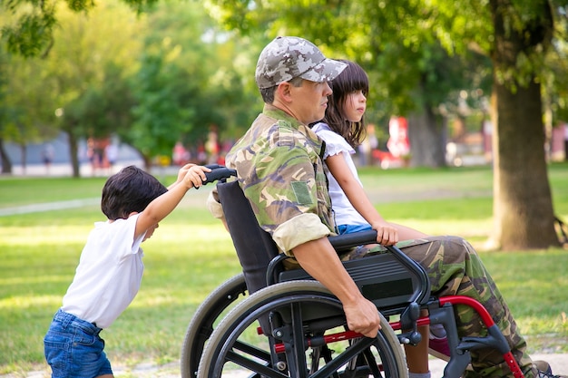 公園で2人の子供と一緒に歩いている障害のある軍のベテラン お父さんの膝の上に座っている女の子 車椅子を押す男の子 戦争または障害の概念のベテラン 無料の写真