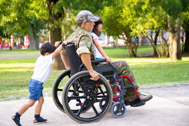 公園で子供たちと一緒に歩いている障害者の引退した軍人 お父さんの膝の上に座っている女の子 車椅子を押す男の子 戦争または障害の概念のベテラン 無料の写真