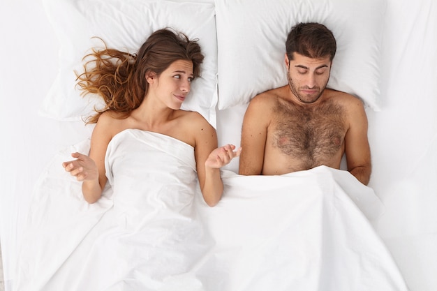 Disappointed man has erectile dysfunction during sex, his woman partner lies near under white blanket, puzzled with husbands impotence, spreads hands sideways. sexual problems. men health concept Free Photo