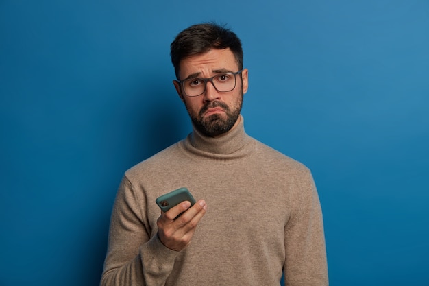 Displeased bearded guy smirks face, uses modern cellphone, has sad expression, wears transparent glasses and jumper Free Photo